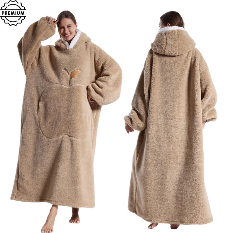 May include: A tan, oversized wearable blanket with a hood and large front pocket. The blanket features an apple design on the front and a white fleece lining around the hood. The blanket is designed to be worn, offering warmth and comfort.