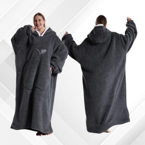 May include: A dark gray oversized wearable blanket with a hood and a large front pocket. The interior of the hood and the cuffs are lined with white fleece. The blanket is worn by a person.