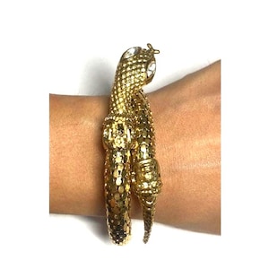 May include: A gold-coloured snake bracelet with a textured, scale-like design. The snake's head features two clear, round gemstones for eyes. The bracelet is wrapped around a wrist.