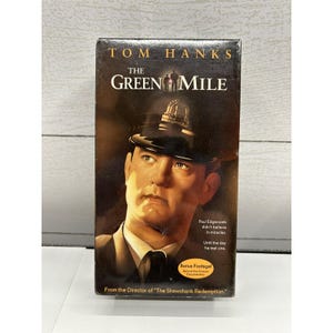 Könnte beinhalten: Eine VHS-Kassette des Films "The Green Mile" mit Tom Hanks. Das Cover zeigt eine Nahaufnahme von Hanks mit einer Polizeimütze. Der Titel ist in großen, fetten Buchstaben gehalten, mit zusätzlichem Text.
