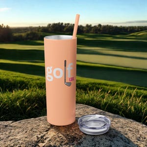 May include: A peach-colored tumbler with a straw and a clear lid. The tumbler has the words "golf girl" in white with a golf club graphic. The background shows a green golf course.