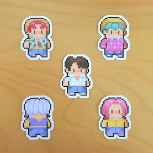 Może przedstawiać: Pięć naklejek w stylu pixel art przedstawiających postacie z kreskówek z różnymi fryzurami i strojami. Postacie są w różnych kolorach, w tym różowym, niebieskim i brązowym. Naklejki znajdują się na jasnobrązowej drewnianej powierzchni.