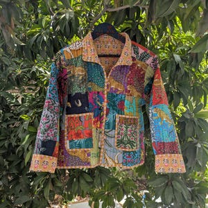 Może przedstawiać: Kurtka patchwork z różnorodnymi, kolorowymi wzorami, w tym kwiatowymi, lampartami i geometrycznymi. Kurtka ma kołnierz, długie rękawy i dwie przednie kieszenie. Wisi na czarnym wieszaku na tle zielonych liści.