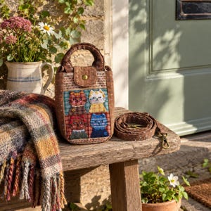 Puede incluir: Un bolso de tela marrón con textura acolchada y un asa superior. El panel frontal muestra cuatro caras de gato bordadas en un patrón de cuadrícula, cada una con colores y expresiones distintas. Se incluye una correa de hombro desmontable a juego.