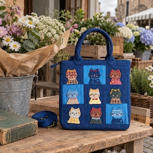 Puede incluir: Un bolso tote acolchado de color azul oscuro con un patrón de cuadrícula de nueve parches bordados de gatos con diversos colores y atuendos. El bolso tiene dos asas superiores y una correa de hombro a juego con herrajes metálicos dorados.