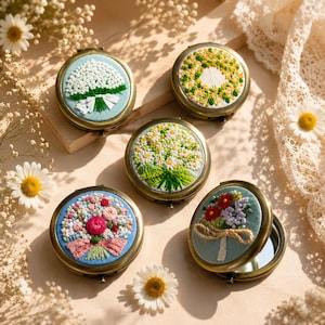 Op de afbeelding: Vijf ronde metalen compactspiegels met geborduurde bloemmotieven op de deksels. Het borduurwerk toont diverse boeketten en kransen in witte, gele, roze, rode en groene draden op een lichtblauwe of witte stoffen ondergrond.