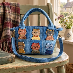 Puede incluir: Un bolso de mano de mezclilla azul con seis parches bordados de gatos en varios colores y patrones. El bolso tiene dos asas superiores y una correa de hombro desmontable sujeta con herrajes de metal en tono latón. Los diseños de gatos muestran diferentes expresiones.