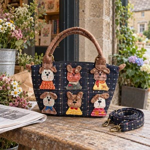 Puede incluir: Un bolso de mano de mezclilla azul oscuro con un patrón de cuadrícula y seis apliques de perros bordados con diversos colores y atuendos. El bolso tiene dos asas texturizadas marrones y una correa de hombro a juego con clips dorados.