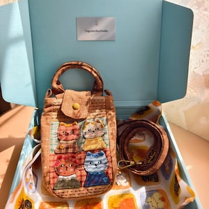 May include: A brown fabric handbag featuring an embroidered grid of four stylized cat faces in red, yellow, orange, and blue. The bag has a top handle, a flap closure with a gold-tone snap, and a detachable brown shoulder strap inside a light blue box.