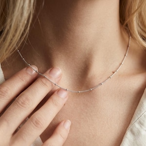 May include: Delicate silver necklace featuring small, evenly spaced beads. The necklace is a simple, elegant design, perfect for everyday wear. The chain is thin and the beads are small, creating a minimalist aesthetic.