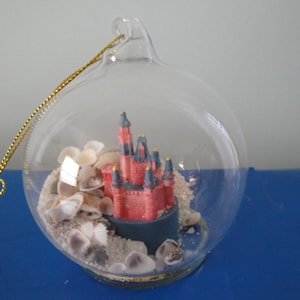 May include: A glass ornament with a miniature red and blue castle inside, surrounded by white seashells and sand.