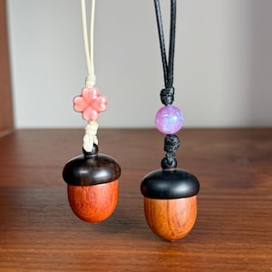 Könnte beinhalten: Zwei eichelförmige Anhänger mit Holzkörpern und dunklen Kappen. Einer hat eine rosa Blumenperle, der andere eine lila Perle. Beide sind an Kordeln aufgehängt und zeigen die natürliche Holzmaserung.