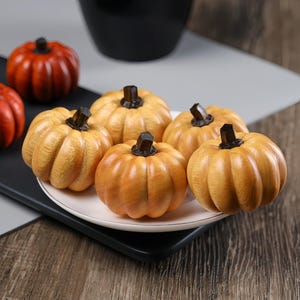 May include: A collection of small, decorative pumpkins with a light orange hue and dark brown stems. The pumpkins are arranged on a white plate and a black tray, with a wooden surface in the background. These fall-themed decorations are perfect for autumn.