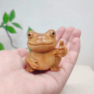 Könnte beinhalten: Eine Holzfroschfigur mit erhobenem Daumen. Der Frosch hat einen lächelnden Gesichtsausdruck und schwarze Augen. Die Figur ist aus hellfarbenem Holz gefertigt und ein dekorativer Gegenstand.