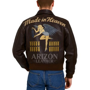 Könnte beinhalten: Dunkelbraune Lederjacke im Bomberstil. Auf der Rückseite der Jacke befindet sich eine Grafik einer Frau mit Engelsflügeln, die eine Bombe hält, mit dem Text "Made in Heaven" und "Arizon Leather".