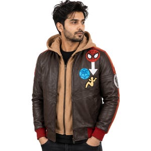 May include: A dark brown leather bomber jacket featuring a tan hooded inner layer, red ribbed cuffs, and various colorful patches on the chest and sleeves, including a Spider-Man mask, a blue web icon, and a yellow figure graphic.