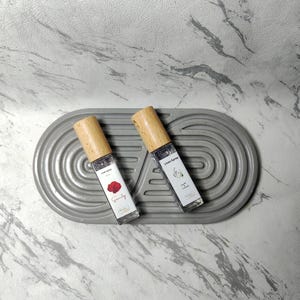 May include: Two glass linen spray bottles with wooden caps rest on a gray, oval-shaped tray. One bottle features a red rose graphic and the word "Serenity." The other displays a lavender and chamomile graphic. Both bottles are labeled "Linen Spray."
