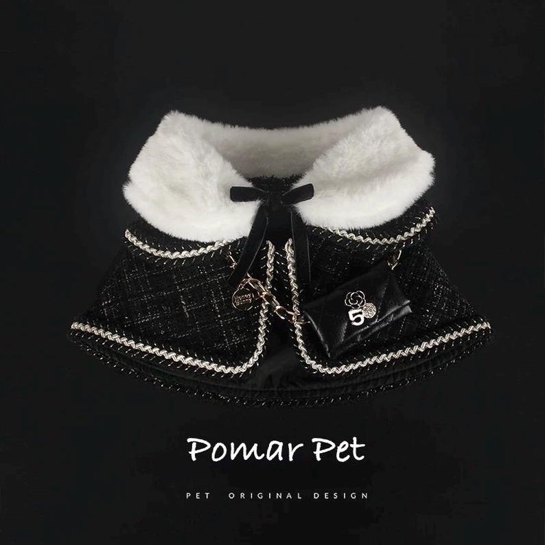 May include: A black and white dog cape with a faux fur collar and a small black purse. The cape has a quilted pattern with white trim and a black bow. The purse has a gold chain and the number 5.
