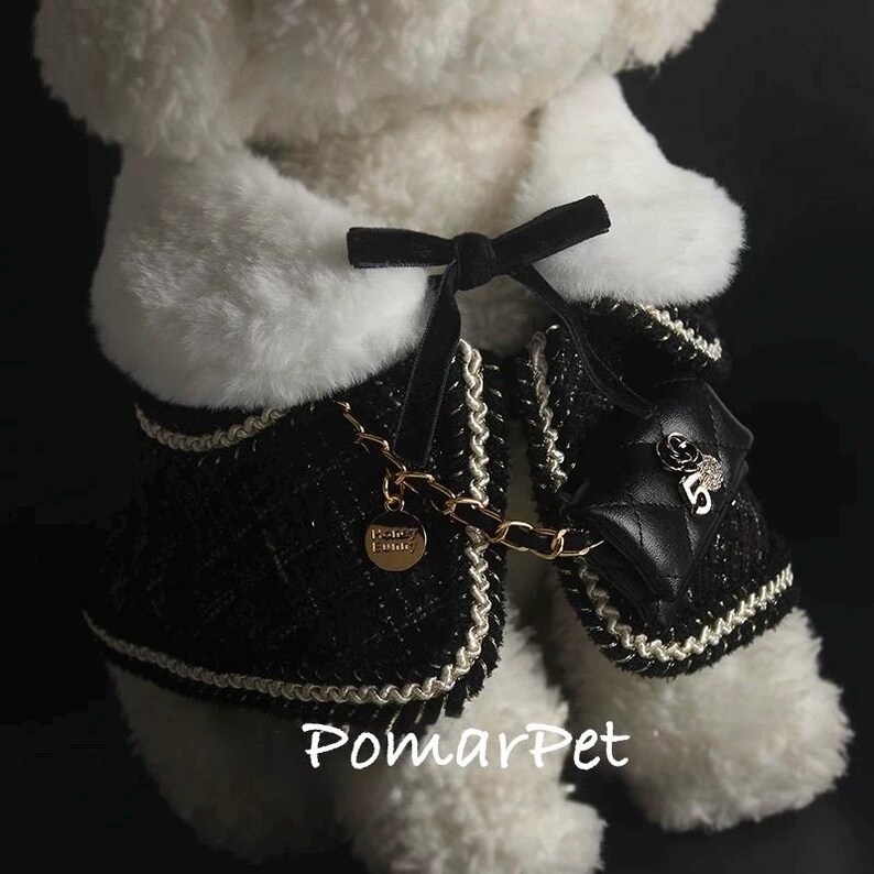May include: A black and white dog jacket with a quilted design, gold chain, and a small purse. The jacket has a black bow and white trim. The text "PomarPet" is visible at the bottom.