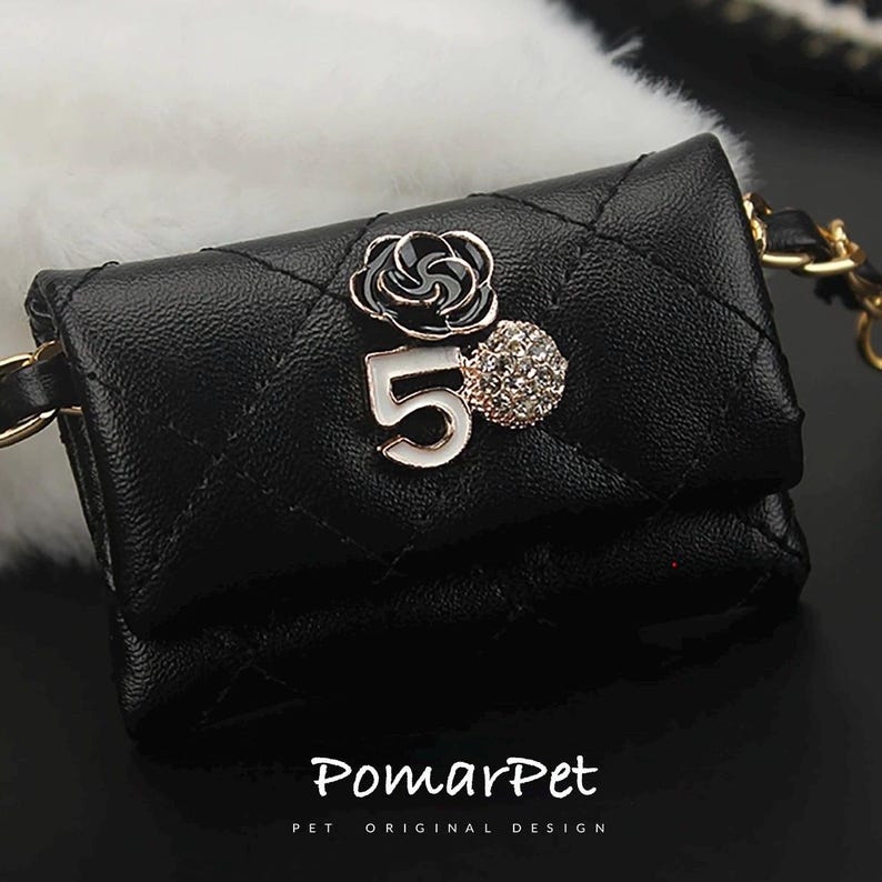 May include: Black quilted pet carrier with a gold chain strap. The carrier features a decorative black and white flower, the number 5, and a rhinestone embellishment. The text "PomarPet" is printed at the bottom.