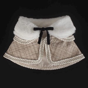 May include: A beige tweed cape with a white faux fur collar and a black velvet bow. The cape is trimmed with a silver braided trim. The garment is designed with a layered effect and is set against a black background.