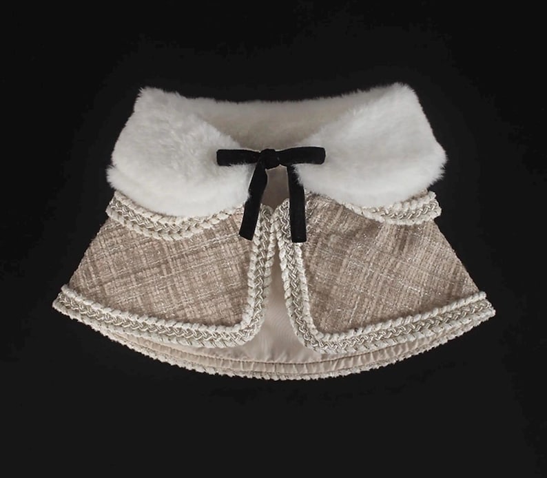May include: A beige tweed cape with a white faux fur collar and a black velvet bow. The cape is trimmed with a silver braided trim. The garment is designed with a layered effect and is set against a black background.