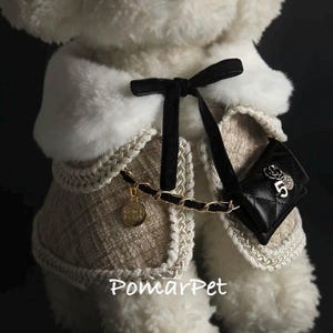 May include: A dog coat in beige and white, featuring a black bow, a small black purse, and a gold chain. The coat has a faux fur collar and gold trim. The purse has a flower and the number 5. The text "PomarPet" is visible.