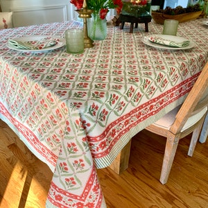 May include: A white tablecloth with a repeating pattern of red flowers and green leaves. The tablecloth has a red border with a striped edge. The table is set with plates, napkins, and glasses. A vase of red flowers and a candle holder are also on the table.
