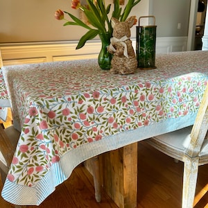 May include: A rectangular tablecloth with a floral pattern of pink fruits and green leaves on a white background. The border features blue and white stripes. A vase of tulips, a bunny figurine, and a green lantern are on the table.