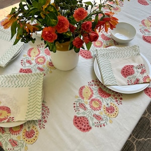 May include: A white tablecloth with a floral pattern in shades of pink, yellow, and green. The tablecloth is set with two place settings, each with a white plate, a white napkin, and a white bowl. A bouquet of orange and yellow flowers is in a white vase in the center of the table.
