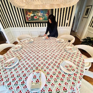 May include: A round dining table set with a floral patterned tablecloth, white plates, and matching napkins. The tablecloth features a red and green floral design on a white background. The table is surrounded by white chairs.