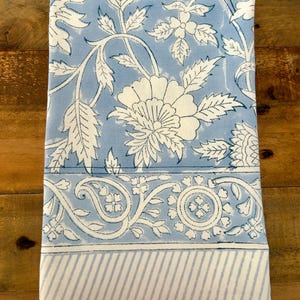 May include: A folded blue and white floral patterned textile. The design features large white flowers and leaves on a light blue background. The bottom border has a striped pattern.