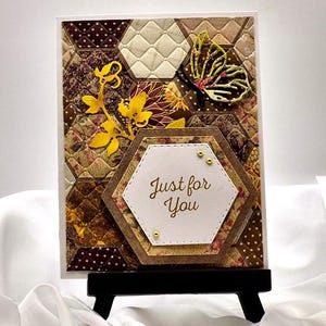 May include: A handmade greeting card with a hexagonal pattern in shades of brown, beige, and yellow. A gold butterfly and floral accents adorn the card. The text "Just for You" is displayed on a white hexagon. The card is displayed on a black easel.