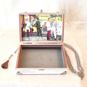 May include: A wooden box purse with a hinged lid, featuring a vintage illustration of people in a shop. The box is trimmed with red and has a silver chain strap and a tassel. The text "ALADINO CONNECTICUT" is visible.