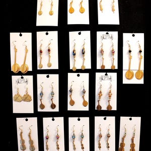 May include: A collection of gold-toned earrings displayed on white cards. The earrings feature various musical instrument designs, including banjos, mandolins, and violins. Some earrings incorporate colorful beads. The background is black.