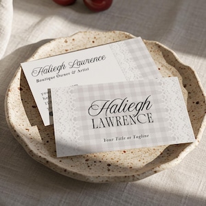 May include: Two business cards with the name "Haliegh Lawrence" and the tagline "Your Title or Tagline" are displayed on a speckled ceramic plate. The cards have a gingham pattern and lace trim. Two cherries sit nearby.