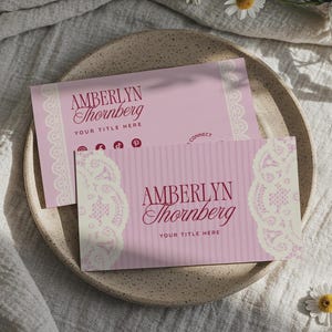 Pode incluir: Dois cartões de visita cor-de-rosa com o texto "Amberlyn Thornberg" e "Your Title Here" são exibidos num prato salpicado. Os cartões têm um desenho de renda no lado esquerdo, sobre um fundo de tecido branco.