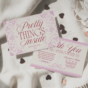 May include: Two pink and white business cards with the text "Pretty Things Inside" and "Thank You for Shopping With Us". The cards have a decorative lace design and are scattered on a white cloth with heart-shaped confetti.