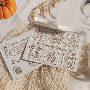May include: A still-life image featuring autumn-themed stationery and pumpkins. The stationery includes cards with fall-themed illustrations and text, alongside a small orange pumpkin and a white pumpkin. A glass of water is also present.