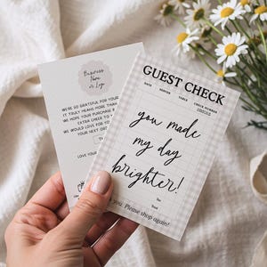Puede incluir: Dos tarjetas impresas se sostienen en una mano, una con un logotipo floral y texto, la otra un cheque de invitado con las palabras "you made my day brighter!". Un ramo de margaritas descansa en el fondo.