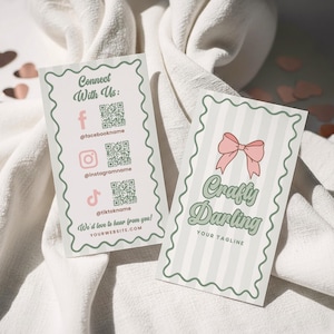 Może przedstawiać: Dwie wizytówki z falistą krawędzią i jasnozieloną obwódką. Jedna karta ma różową kokardę i napis "Crafty Darling". Druga karta zawiera ikony mediów społecznościowych i tekst "Połącz się z nami". Obie karty leżą na białej tkaninie.
