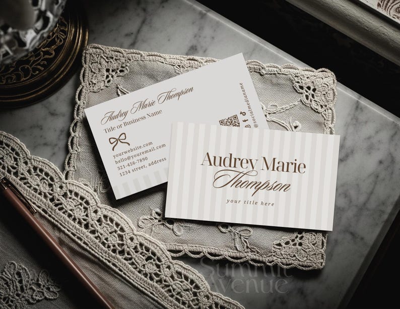 Puede incluir: Dos tarjetas de visita con el nombre "Audrey Marie Thompson" en escritura elegante. Una tarjeta presenta un lazo y detalles de contacto, la otra tiene rayas verticales. Ambas tarjetas descansan sobre un encaje.
