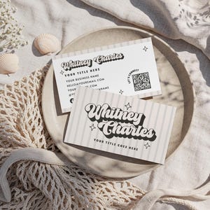 May include: Two business cards with the text "Whitney Charles" in a retro font, with star accents. One card has a QR code and contact information. The cards are on a neutral-toned plate, with seashells and a net bag.