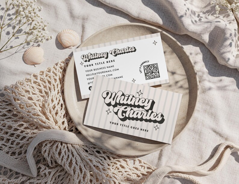May include: Two business cards with the text "Whitney Charles" in a retro font, with star accents. One card has a QR code and contact information. The cards are on a neutral-toned plate, with seashells and a net bag.