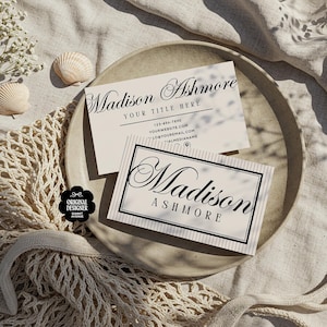 May include: Two business cards with the name "Madison Ashmore" in elegant script are displayed on a beige plate. One card has a striped background. The plate rests on a textured beige surface, with seashells and flowers nearby.