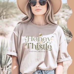 May include: A woman wearing a beige t-shirt with the text "Honey Thistle Custom Creations" and a bee graphic. She is wearing a wide-brimmed hat and sunglasses.
