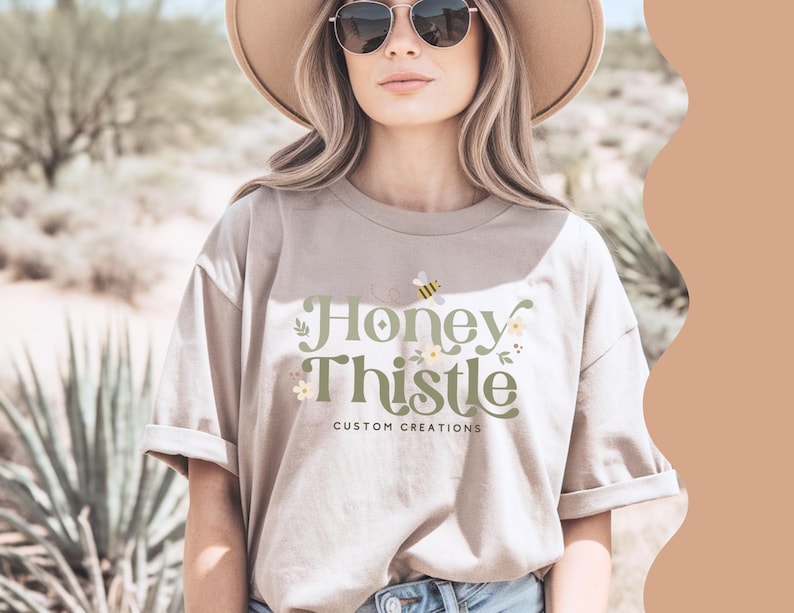 May include: A woman wearing a beige t-shirt with the text "Honey Thistle Custom Creations" and a bee graphic. She is wearing a wide-brimmed hat and sunglasses.