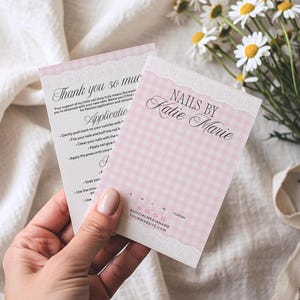 Op de afbeelding: Twee roze en witte visitekaartjes met de tekst "NAILS BY Katie Marie" en "Thank you so much". De kaarten hebben een ruitpatroon en een kanten rand. Een hand houdt de kaarten vast, met madeliefjes op de achtergrond.