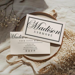 May include: Two business cards with the name "Madison Ashmore" in elegant script. One card is on top of the other, with the top card featuring a striped background and a black border. The bottom card includes contact information. The cards are on a plate.