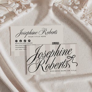 May include: Two business cards with the name "Josephine Roberts" in elegant script. One card has a vertical striped background and the other has social media icons and contact information. The cards are on a white fabric surface.